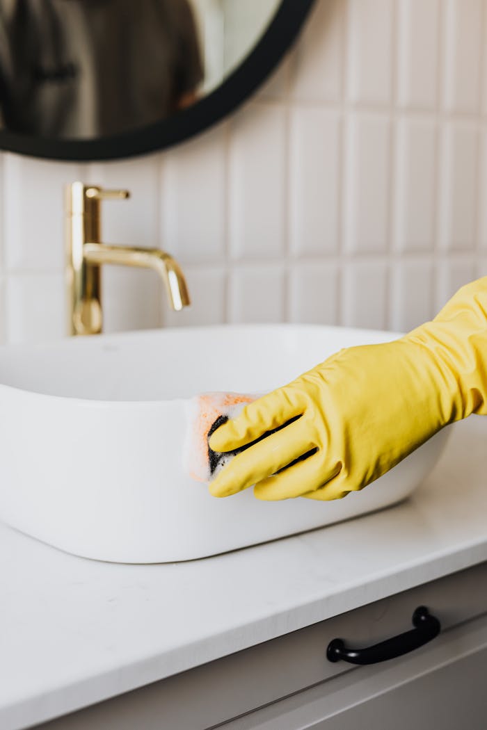 our-services-05 Crop faceless person in yellow rubber protective glove wiping surface of white sink in bathroom with sponge and cleaning agent