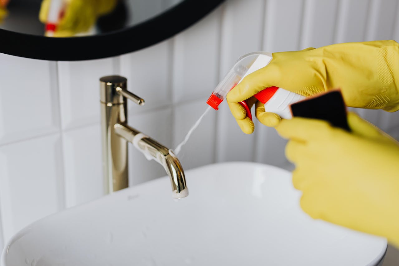 about-img From above anonymous housekeeper in yellow rubber gloves spraying cleaner to chrome water faucet in bathroom